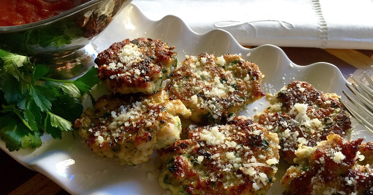 Golden zucchini fritters topped with grated cheese served on a white platter with fresh parsley.