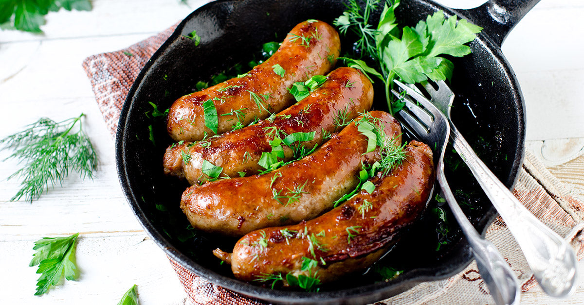 Skillet of browned sausages garnished with fresh herbs and served with utensils.