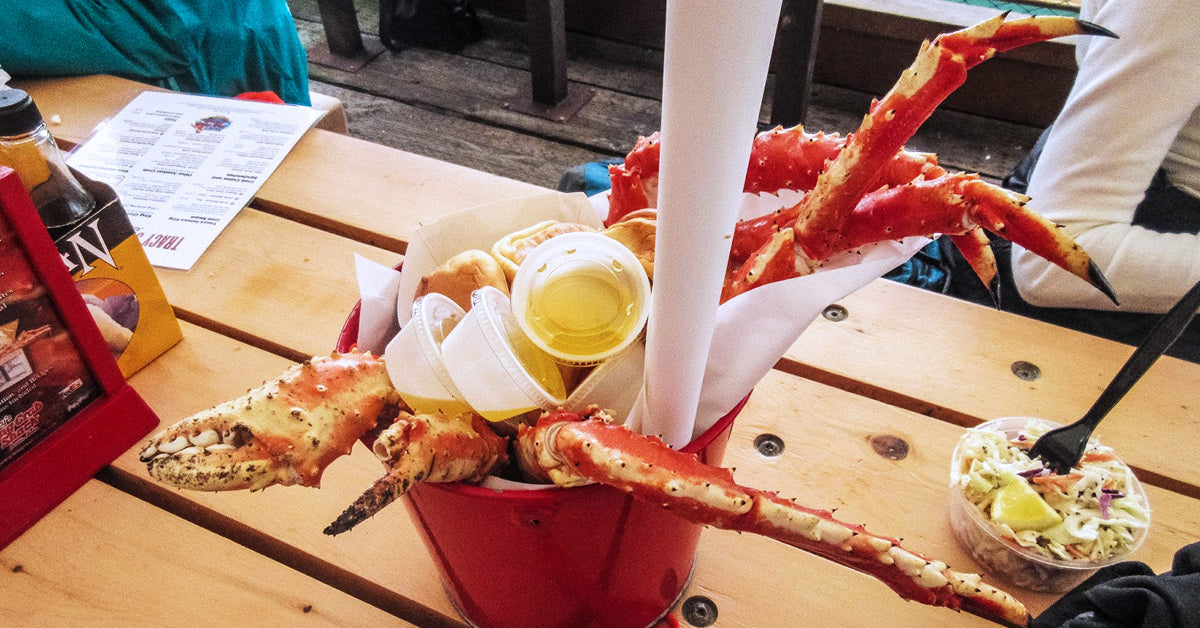 Bucket filled with large crab legs, butter cups, and rolls on a wooden table beside coleslaw and condiments.
