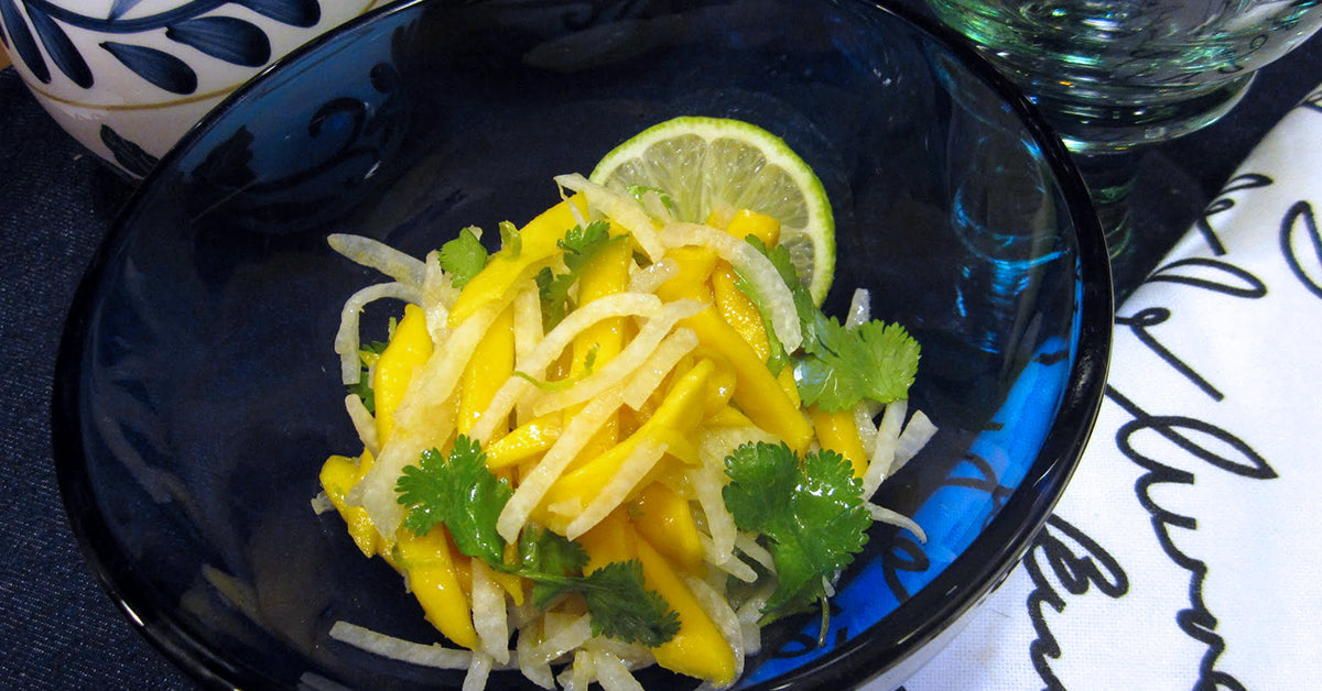 A blue bowl holds a vibrant salad of sliced mango, jicama, and cilantro, garnished with a lime wedge, set against a dark tablecloth and decorative glassware in the background.