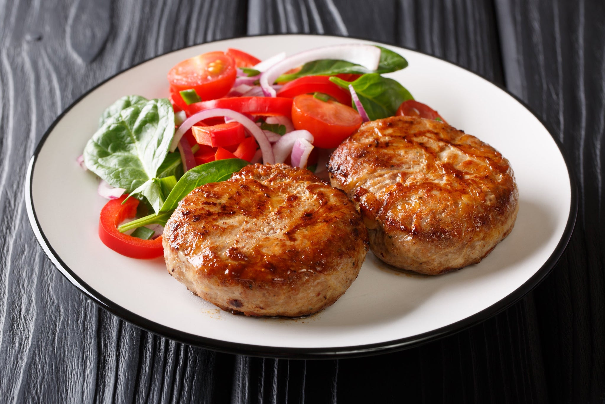 Plate with two grilled meat patties served alongside a fresh salad of tomatoes, spinach, peppers and onions.