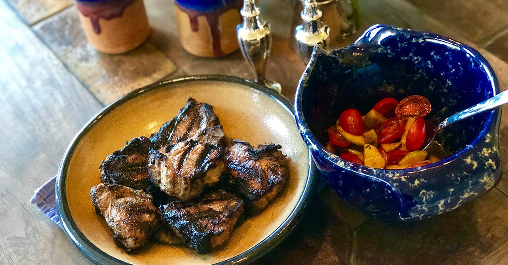 Grilled meat pieces on a plate beside a blue bowl filled with cherry tomatoes and sliced vegetables.