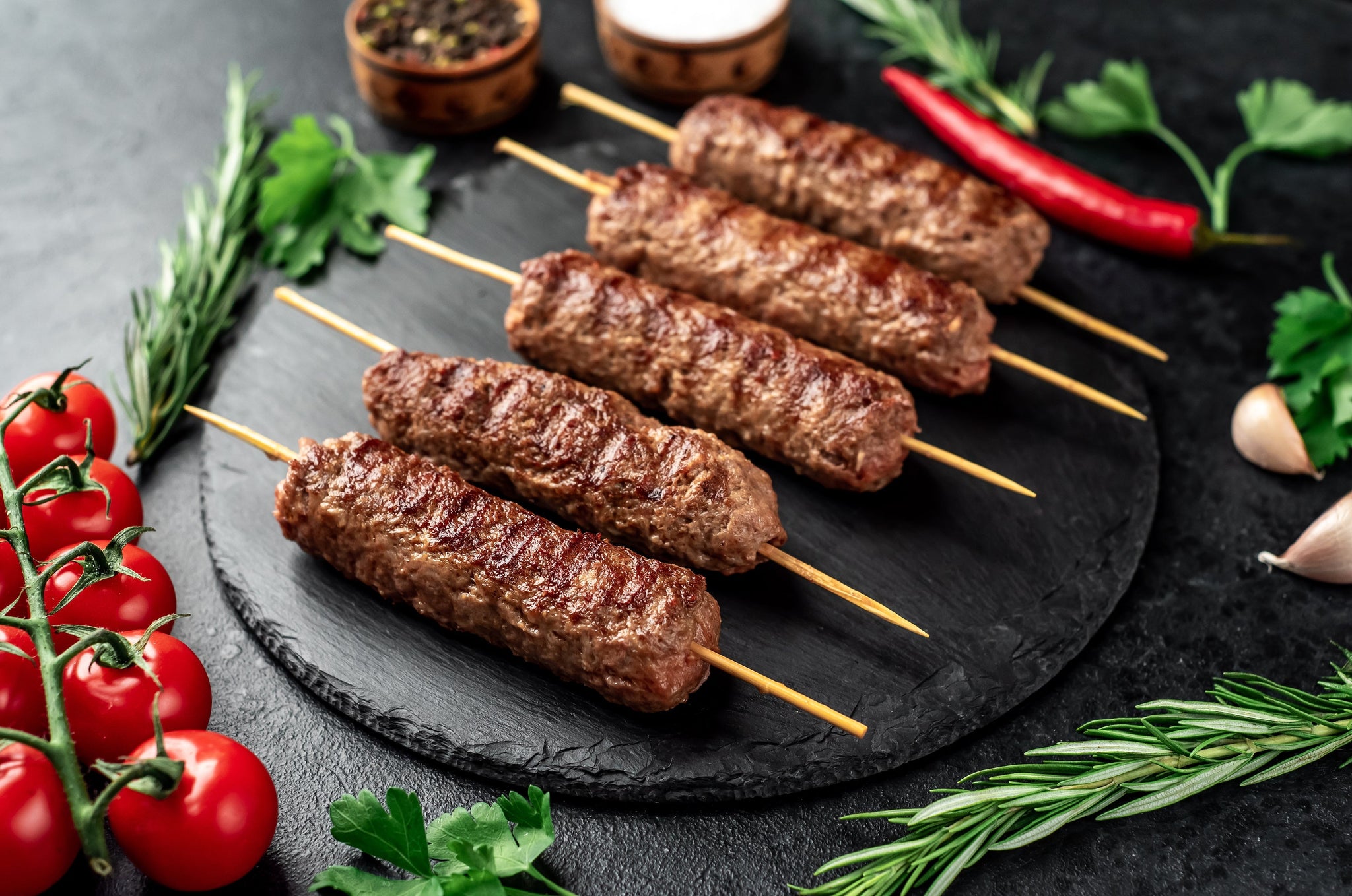 Grilled minced meat skewers arranged on a black slate plate with tomatoes, herbs, garlic, and chili around them.