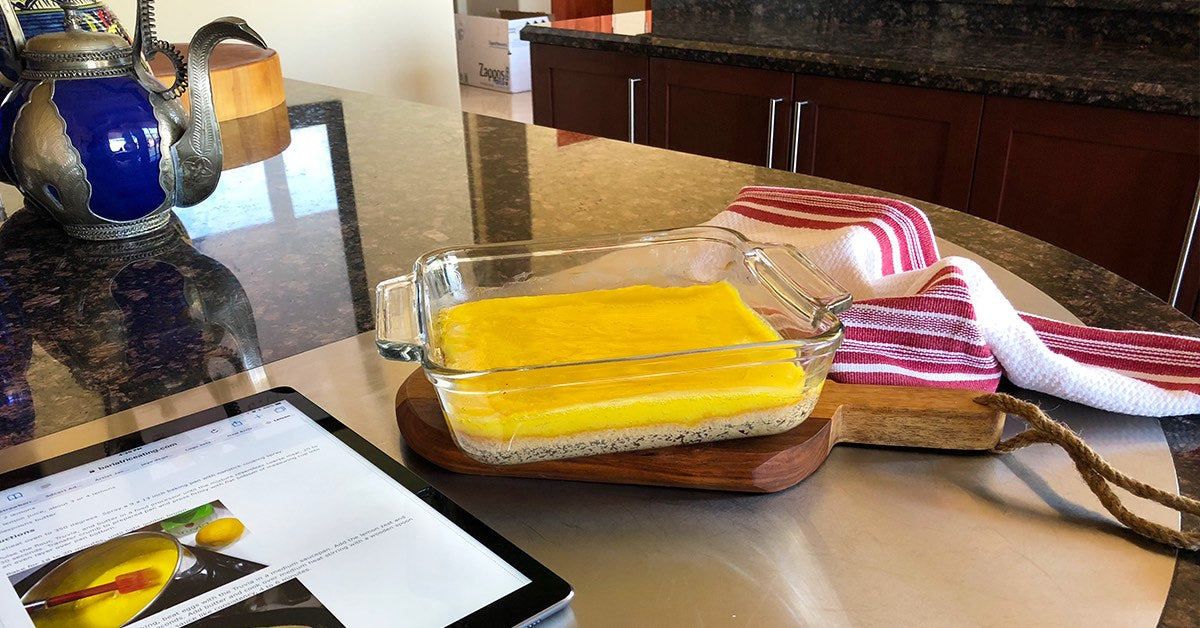 Glass dish of layered yellow dessert on a cutting board beside a tablet and striped kitchen towel on a countertop.