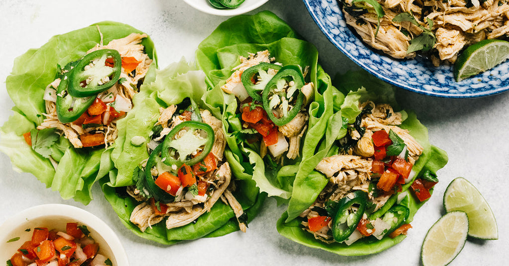 Lettuce wraps filled with shredded chicken, diced tomatoes, and jalapeño slices are arranged on a light surface, accompanied by a bowl of salsa and lime wedges.