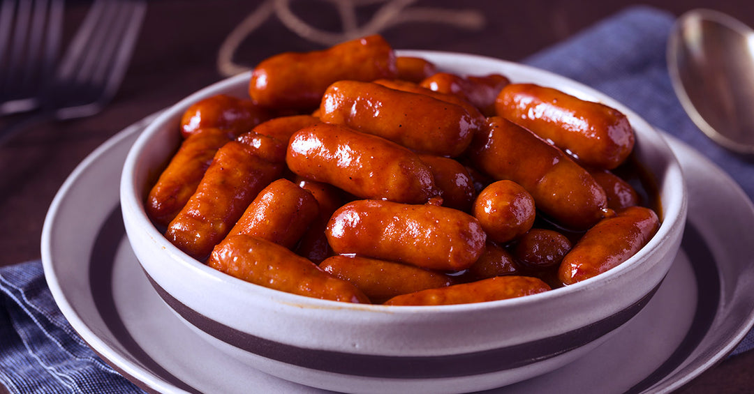 Bowl of glazed cocktail sausages coated in a shiny, reddish-brown sauce on a table setting.