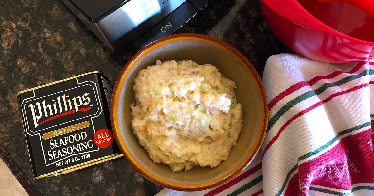 Bowl of creamy seafood mixture beside a can of Phillips seafood seasoning on a kitchen counter with a towel and utensils.