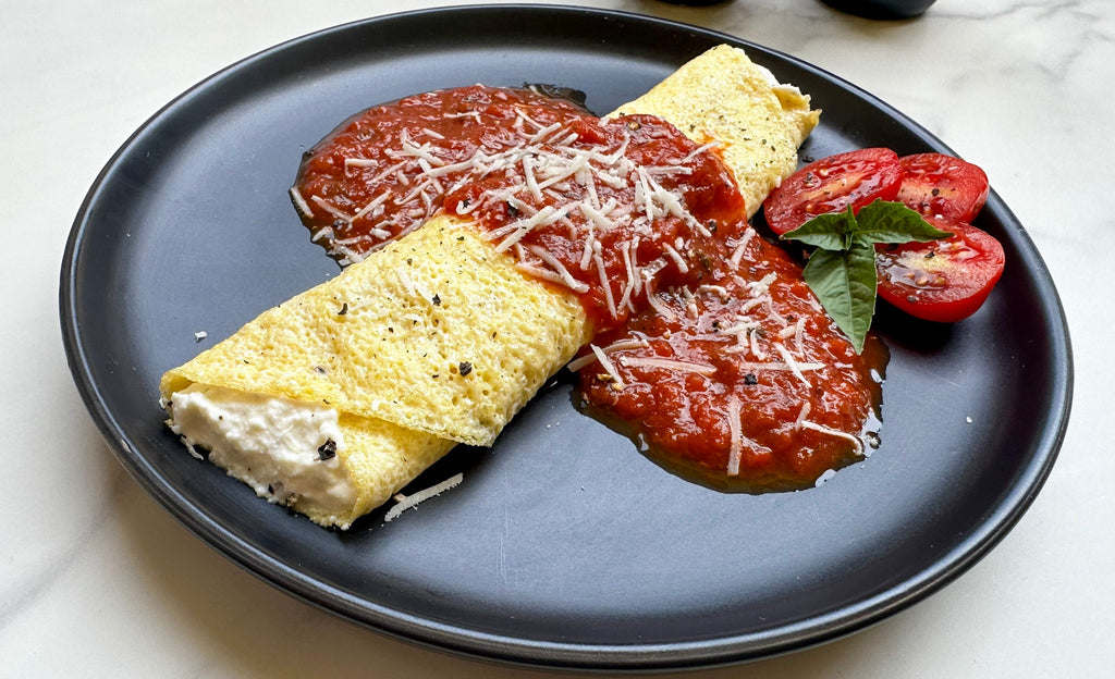 Egg crepe filled with ricotta, topped with marinara and cheese, served with sliced tomatoes and basil on a black plate.