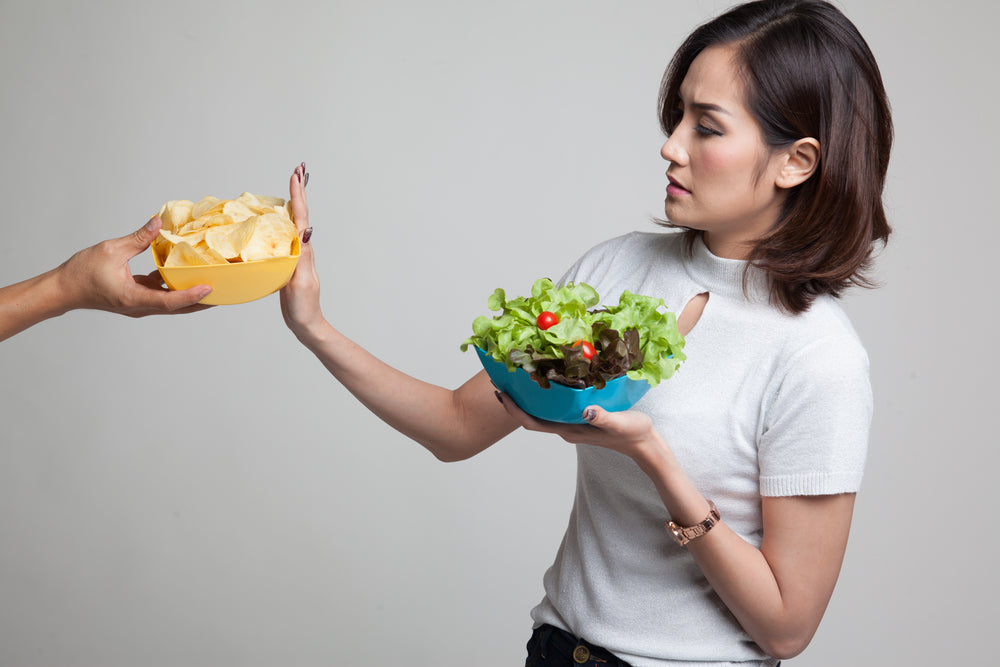 A woman in a light gray top looks disapprovingly at a bowl of chips offered to her while holding a bowl of salad, indicating a choice between unhealthy and healthy food options.