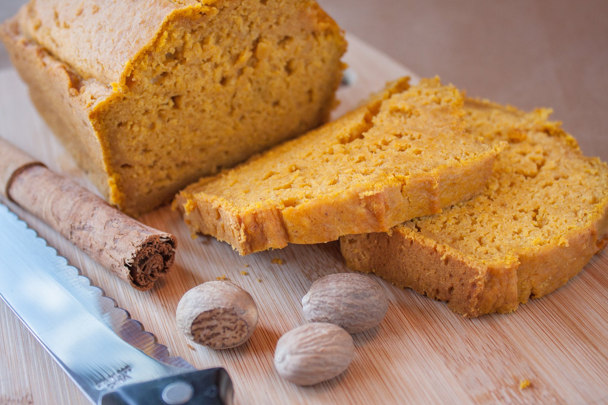 Sliced pumpkin bread on a cutting board with whole nutmeg, a cinnamon stick, and a serrated knife.