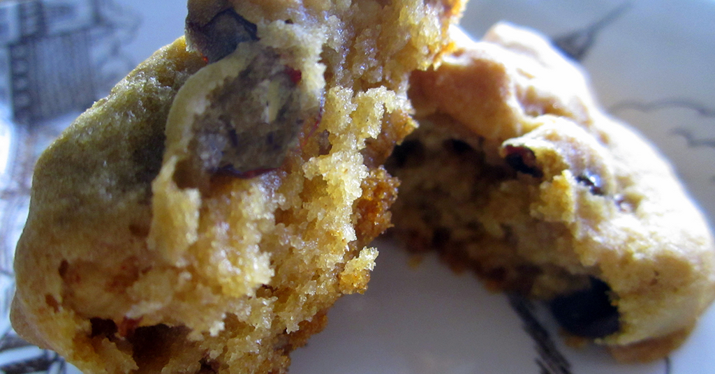 A close-up of a freshly baked chocolate chip cookie, showing a soft, chewy interior with chocolate pieces, resting on a decorative plate with faint patterns.