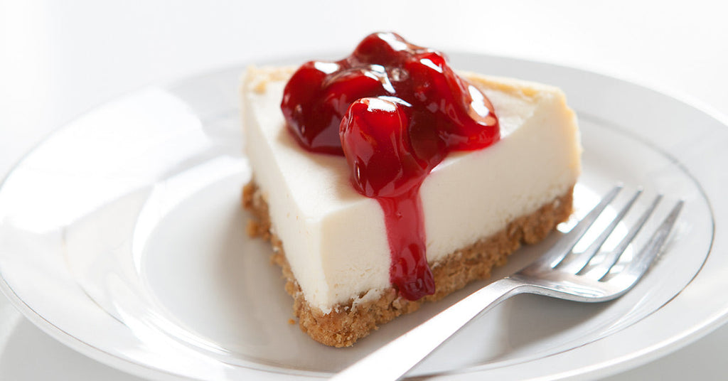 Slice of cheesecake with a graham crust topped with glossy cherry sauce on a white plate with a fork.