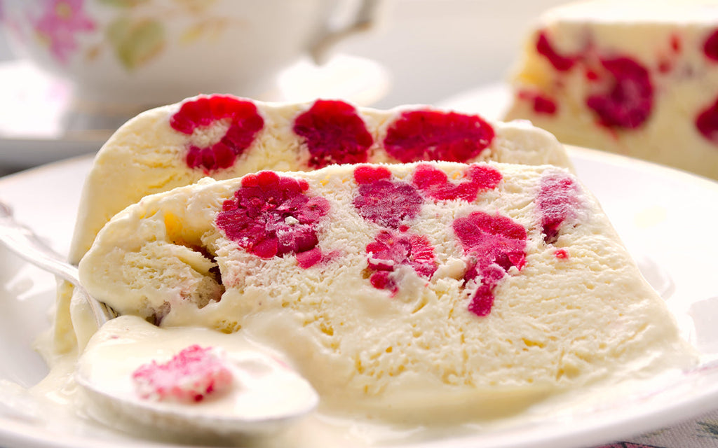 A slice of raspberry ice cream dessert with bright red raspberry swirls is served on a white plate, accompanied by a spoon, with a floral teacup blurred in the background.