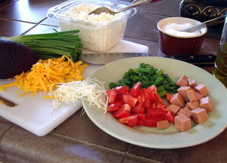 Chopped tomatoes, green onions, cheese, and diced meat arranged on a plate beside grated cheese and avocado on a cutting board.