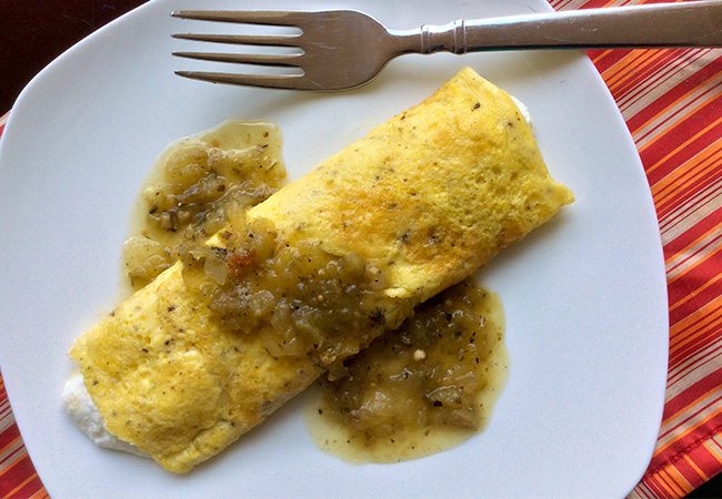 Folded yellow omelet covered with green salsa on a white plate beside a fork on a red and orange striped tablecloth.