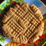 A round, golden-brown peanut butter cookie with a crisscross pattern sits on a colorful plate decorated with fruits and leaves.
