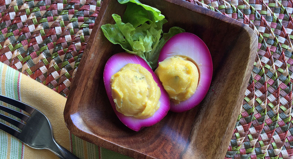 Pink beet-dyed deviled eggs with yellow filling, served with a small bunch of fresh green lettuce on a wooden plate.