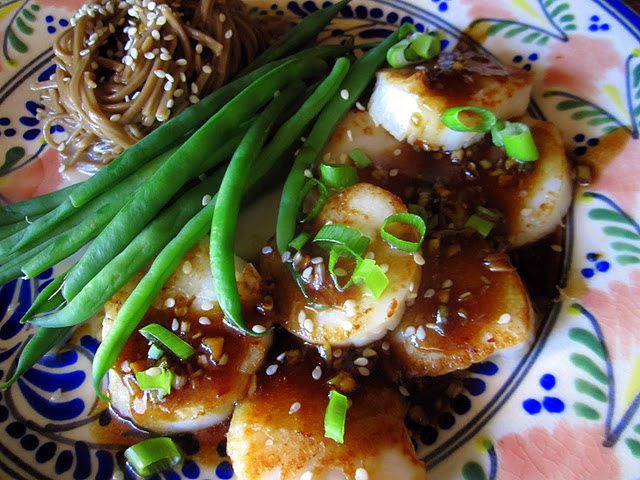 Seared scallops with sauce, topped with green onions, served with green beans and soba noodles on a patterned plate.