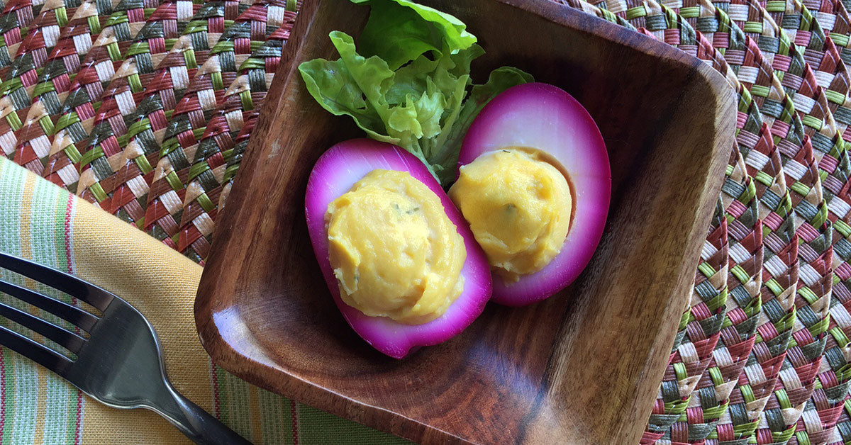 Two deviled eggs with yellow filling are presented in a wooden bowl, surrounded by a piece of lettuce, on a colorful woven placemat next to a fork and a folded napkin.