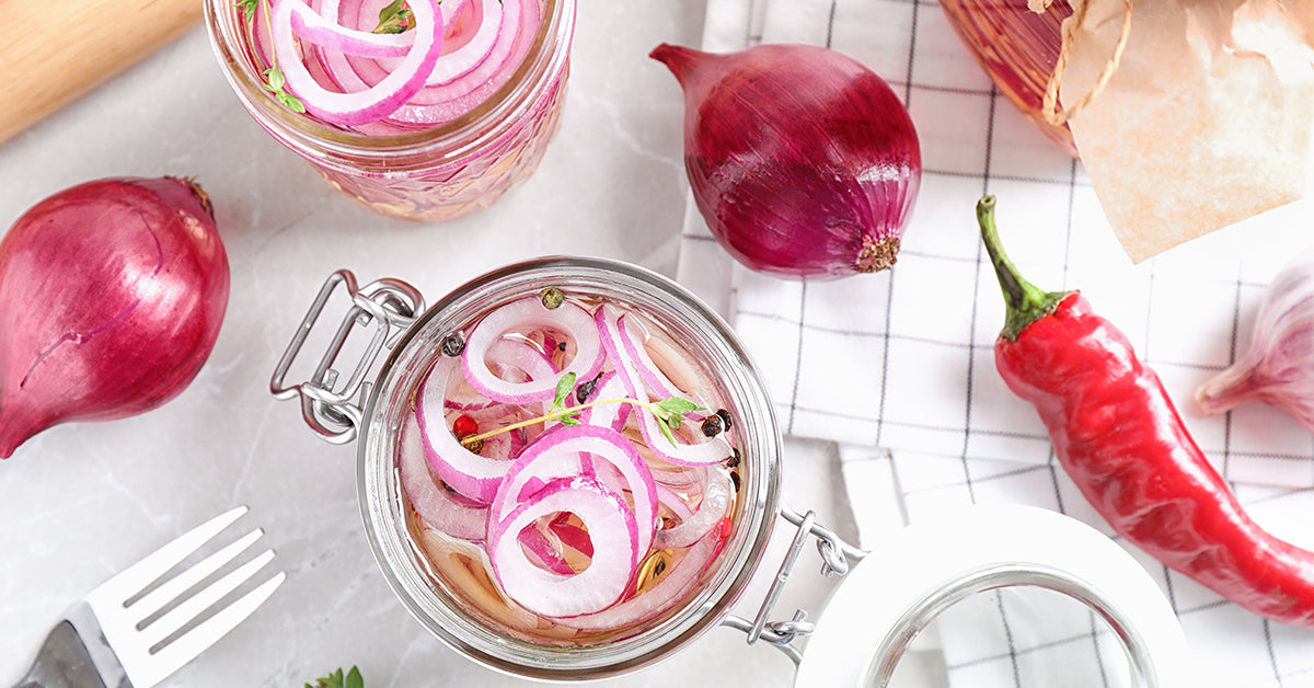 Two glass jars filled with pickled red onions are placed on a marble surface, surrounded by whole red onions, a red chili pepper, garlic, and a fork on a checkered cloth.