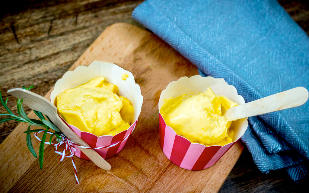 Two cups of yellow ice cream sit on a wooden board, each with a wooden spoon and decorated with a sprig of rosemary, alongside a folded blue cloth.