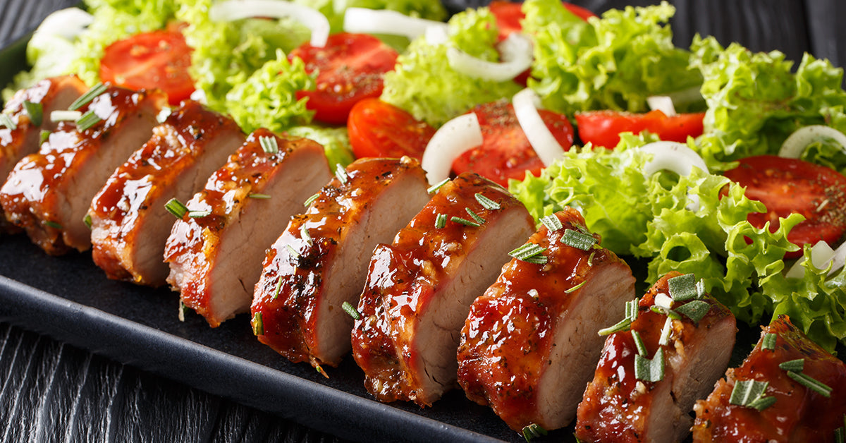 Slices of glazed roasted meat served with fresh lettuce, tomato, and onion salad on a dark plate.