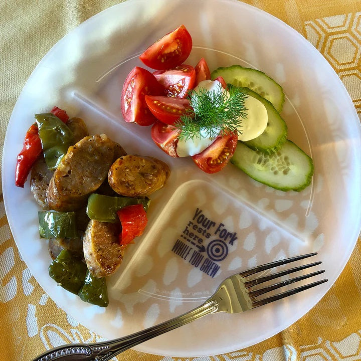 A white plate holds sliced sausage with green and red peppers on one side, and a salad of cherry tomatoes, cucumber slices, and a dollop of sauce on the other, with a fork resting beside it.