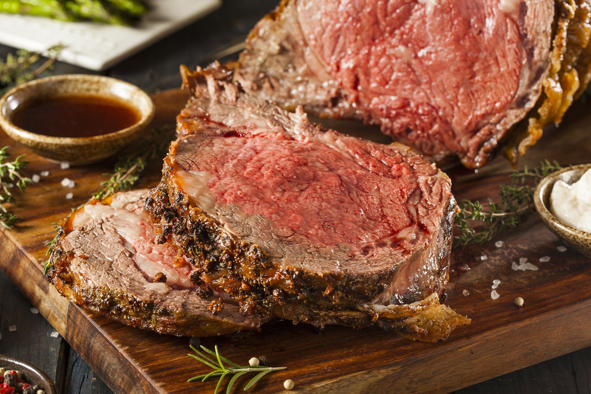 Thick slices of medium-rare roasted beef on a wooden board with herbs, salt, and dipping sauces.