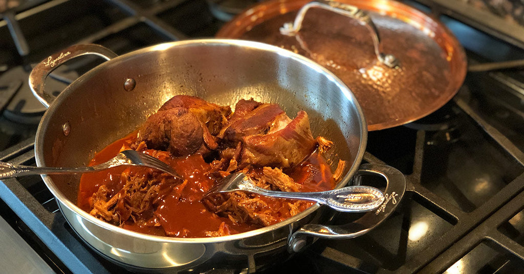 A silver pot on a stovetop contains shredded meat in a thick sauce, with two serving spoons resting inside, and a copper lid partially visible in the background.