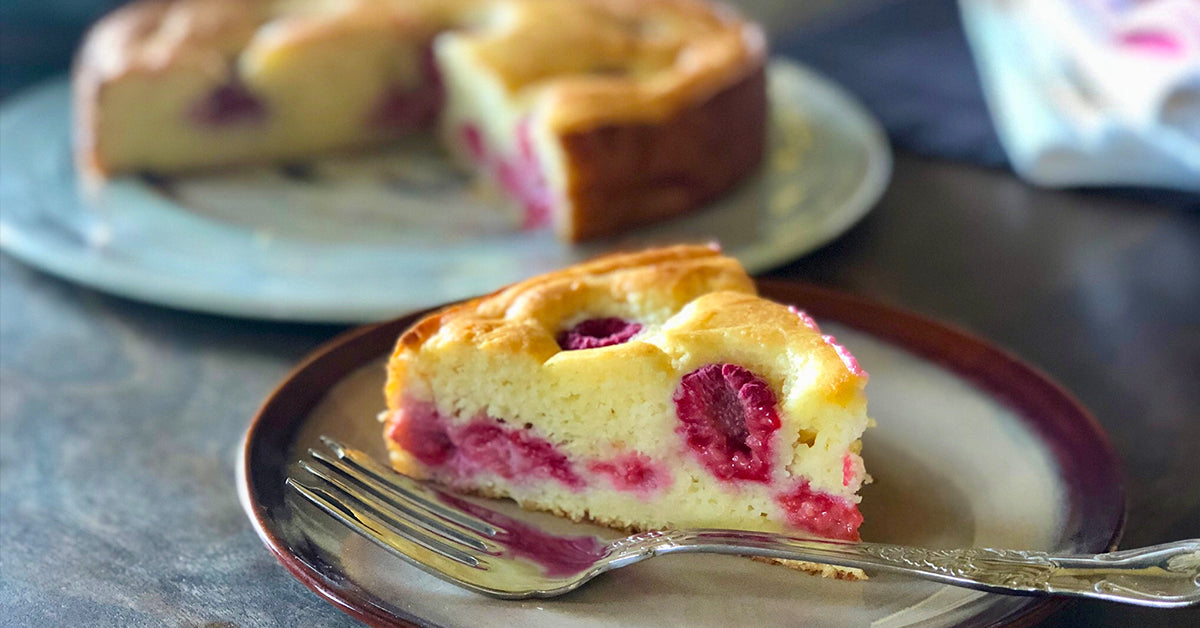 A slice of raspberry ricotta cake sits on a brown plate with a fork beside it, showcasing its moist texture and pink raspberry swirls, while a whole cake is blurred in the background.