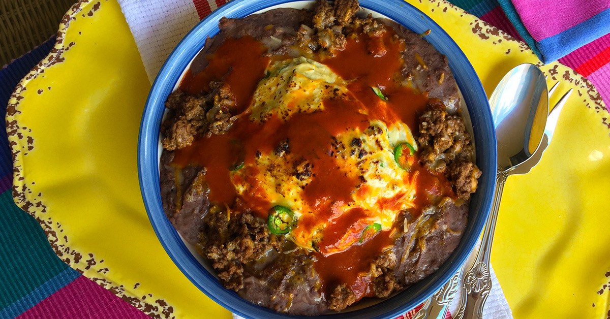 Bowl of refried beans topped with eggs, ground meat, jalapeños, and red sauce on a yellow plate.