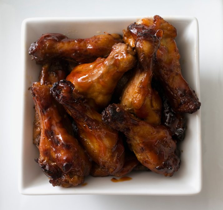 A square white bowl filled with glazed chicken wings, showcasing a mix of golden and dark brown colors, sits against a plain background.