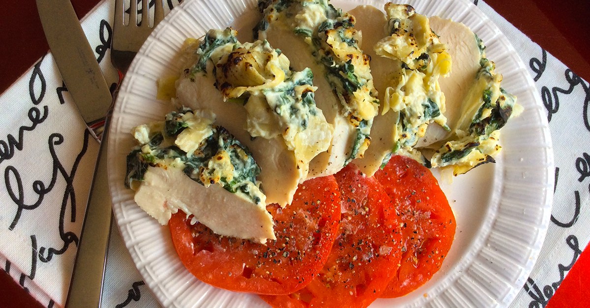 Sliced chicken topped with creamy spinach-artichoke mixture served over peppered tomato slices on a white plate.