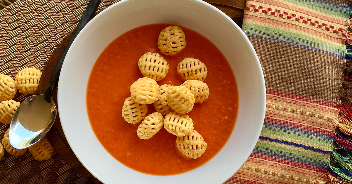 Bowl of tomato soup topped with small lattice-shaped crackers, set on a woven mat beside a colorful striped cloth.