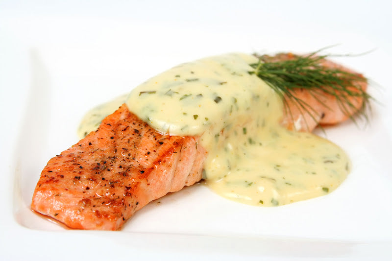 A piece of grilled salmon is topped with a creamy herb sauce and garnished with a sprig of dill, served on a white plate.