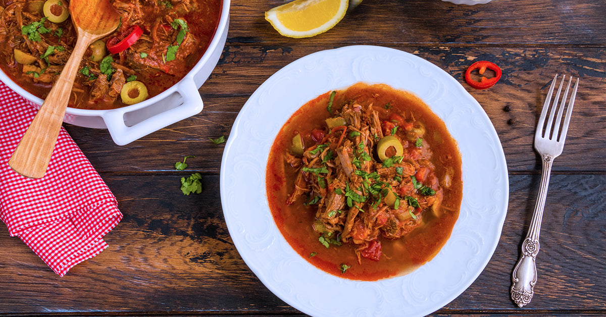 Plate of shredded meat stew with olives and herbs, served beside a pot of the same dish on a wooden table.