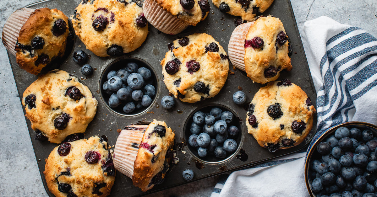 A tray of freshly baked sugar-free blueberry muffins, golden brown with dark blueberries, sits alongside a bowl of blueberries and a striped cloth, showcasing a delicious Sunday morning treat.