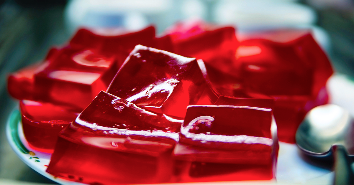 A close-up of shiny red gelatin cubes stacked on a white plate, with a silver spoon nearby, suggesting a sweet treat ready to be served.