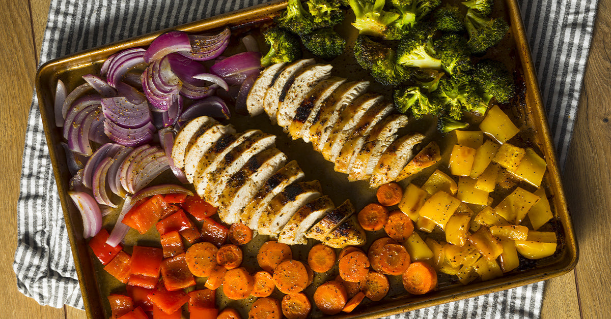 A baking sheet displays sliced grilled chicken breast alongside colorful roasted vegetables, including red onions, bell peppers, broccoli, carrots, and yellow squash, on a striped cloth.