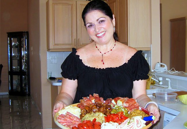 Smiling woman in a kitchen holding a large platter of assorted meats, cheeses, and vegetables.