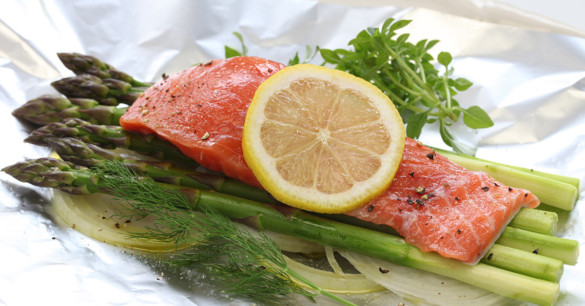 Salmon fillet on asparagus and onions topped with a lemon slice, herbs, and seasoning on foil.