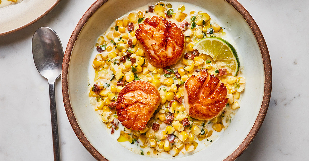 A bowl of creamy corn chowder features three golden-brown seared scallops, garnished with lime, and sprinkled with herbs and diced bacon, set on a light marble surface.
