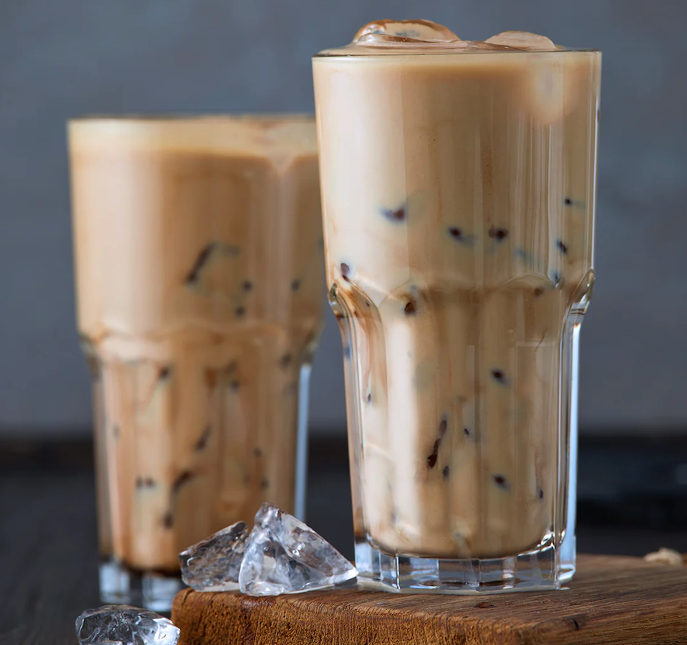 Two tall glasses of iced coffee with ice cubes sit on a wooden surface, showcasing a creamy brown color, with one glass slightly in front of the other against a muted background.