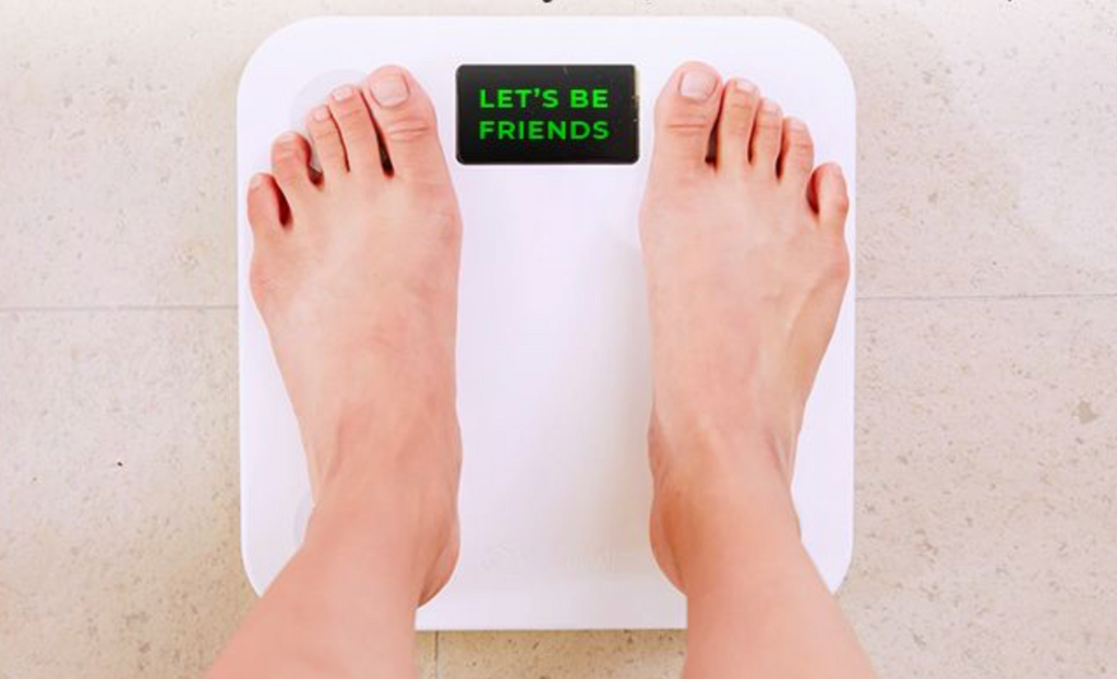 Two bare feet stand on a white digital scale displaying the message "LET'S BE FRIENDS" in bright green letters, set against a light-colored floor.