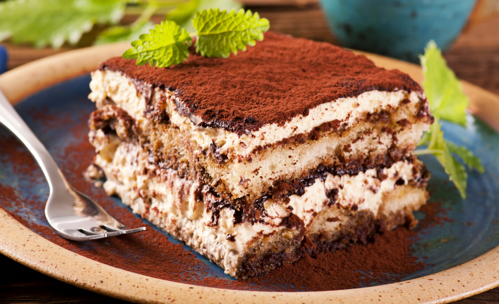 Slice of tiramisu dusted with cocoa powder on a plate, garnished with fresh mint.