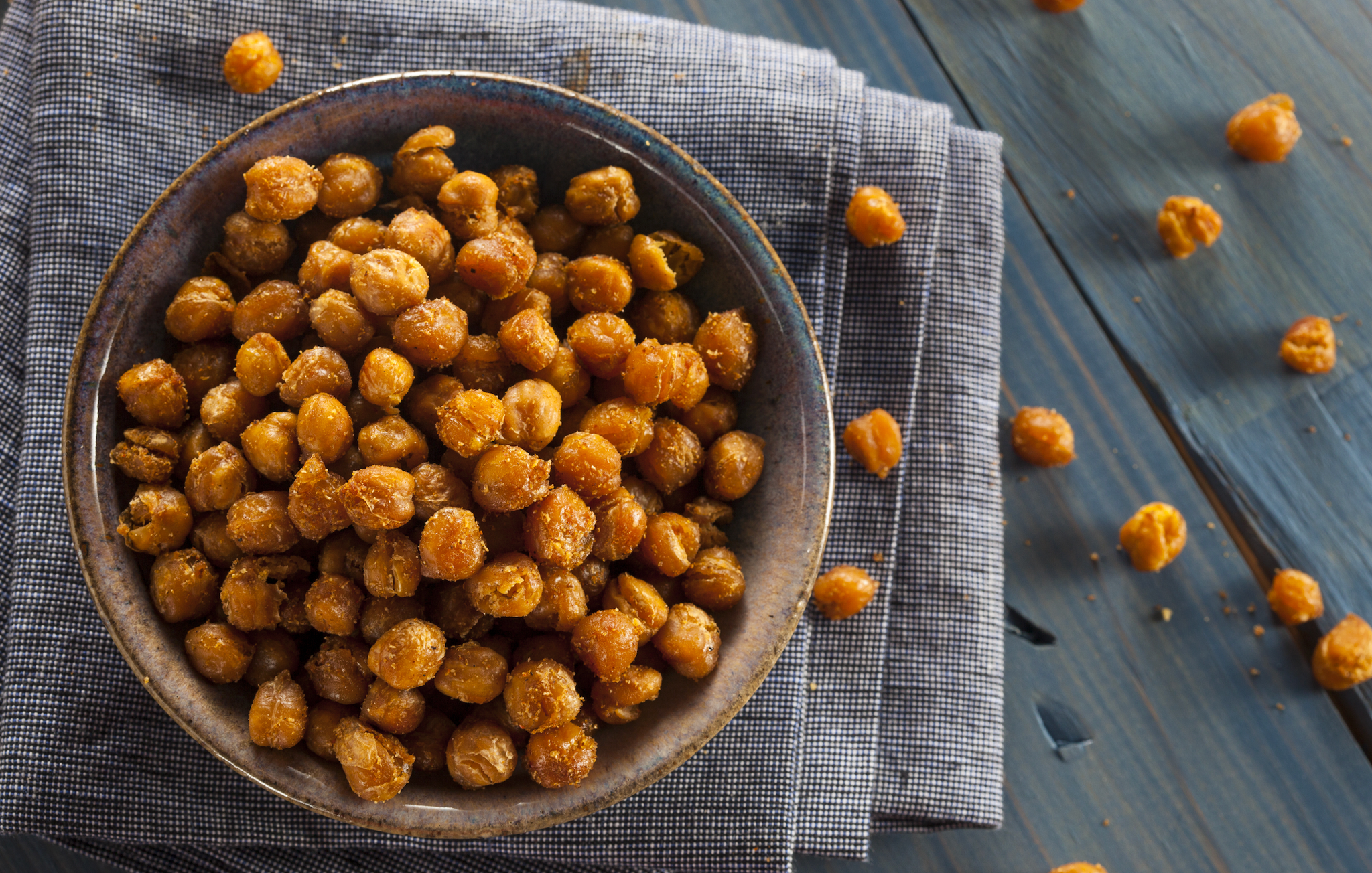 Bowl of crispy roasted chickpeas with scattered pieces on a cloth napkin and wooden surface.