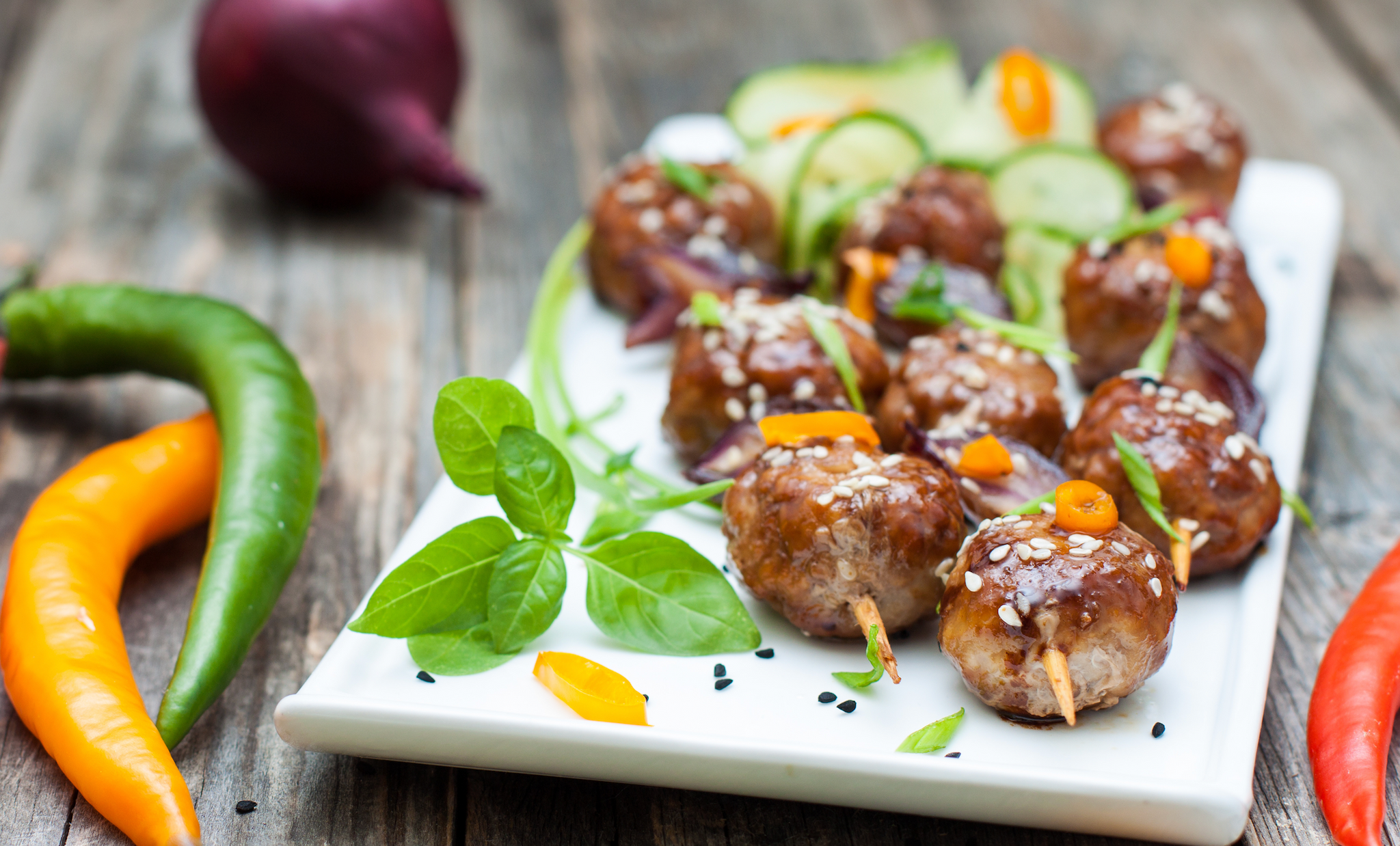 Plate of glazed meatballs topped with sesame seeds and herbs, served with sliced vegetables and peppers.