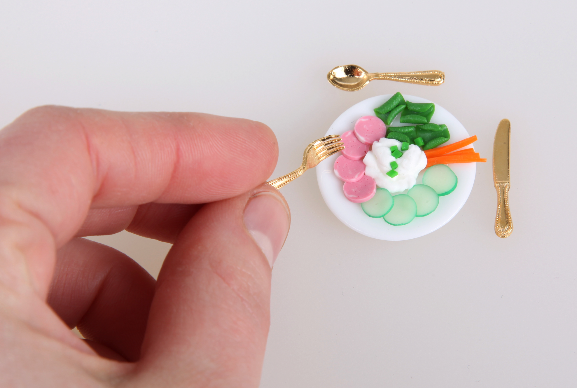Hand holding a tiny fork beside a miniature plate of vegetables and deli slices with tiny gold utensils.