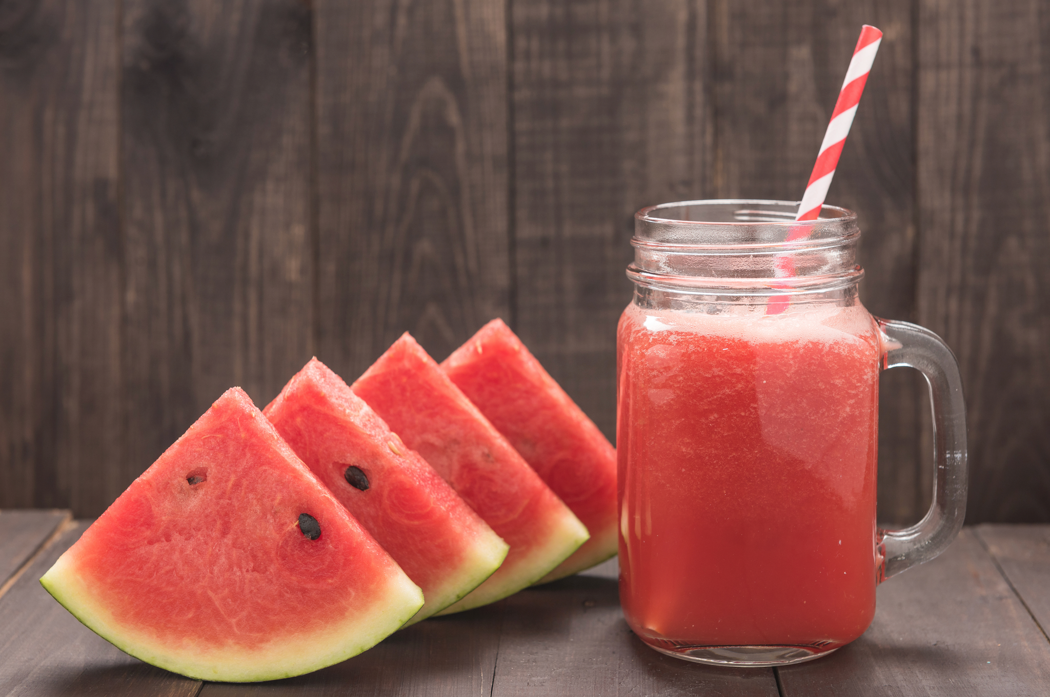 Watermelon slices arranged beside a mason jar filled with watermelon juice and a red-striped straw on a wooden surface.
