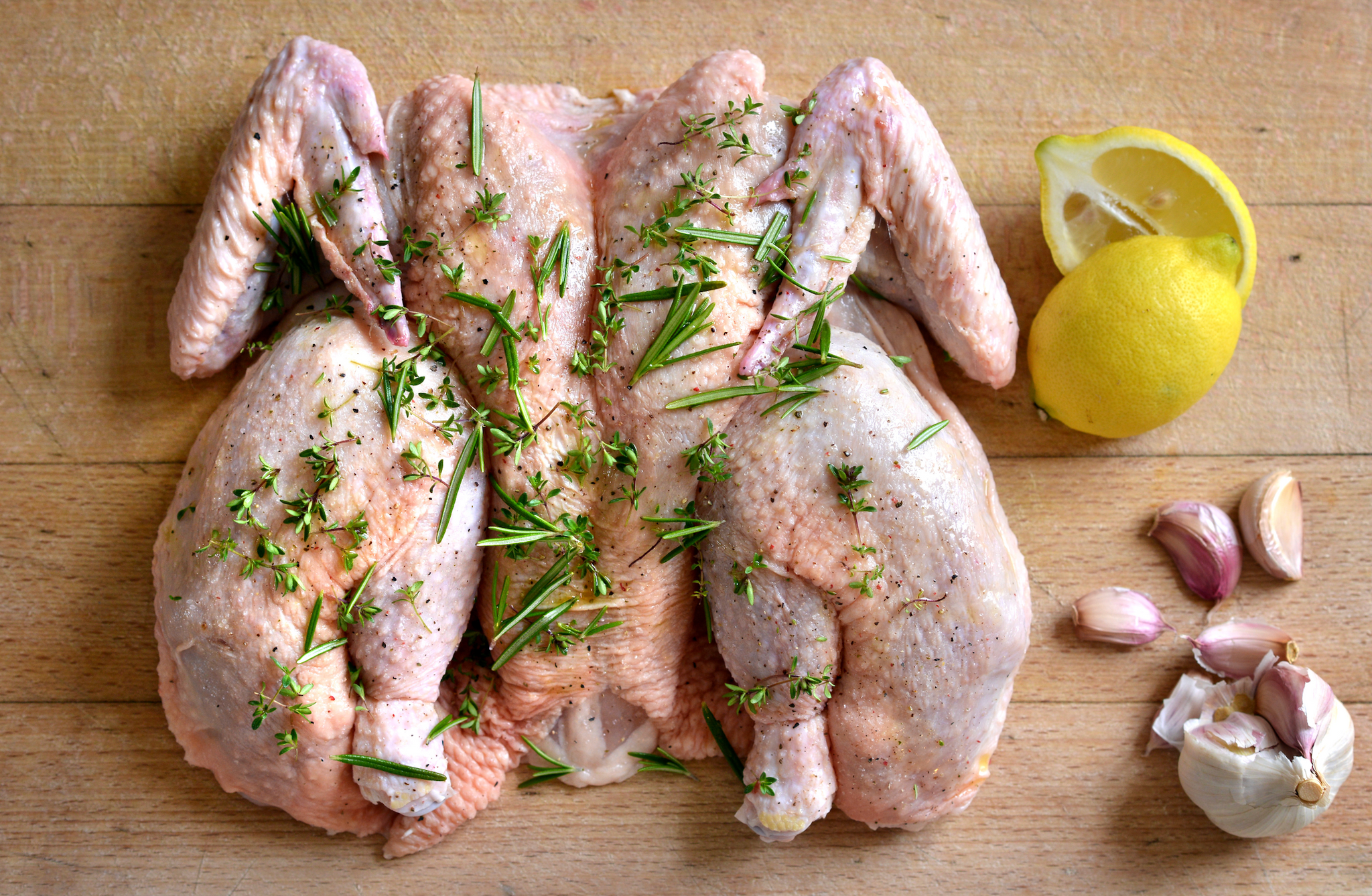 Raw seasoned spatchcocked chicken on a cutting board with fresh herbs, lemon, and garlic.